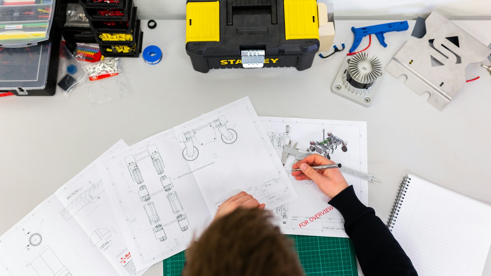 An engineer checking printed technical drawings at a desk with a caliper and pen.